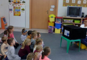 Grupa dzieci siedząca przed telewizorem i nauczycielka przesuwająca taśmę filmową .